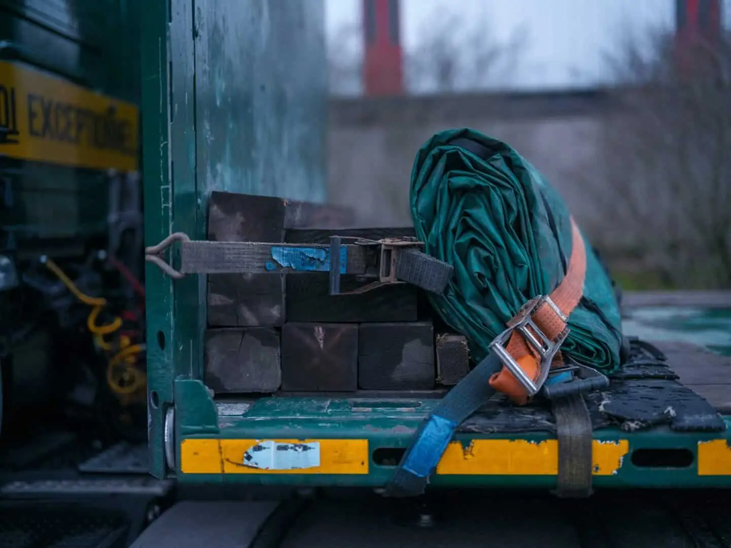 Black timber beams secured with an orange ratchet strap and a rolled green tarpaulin on a flatbed vehicle for safe transit