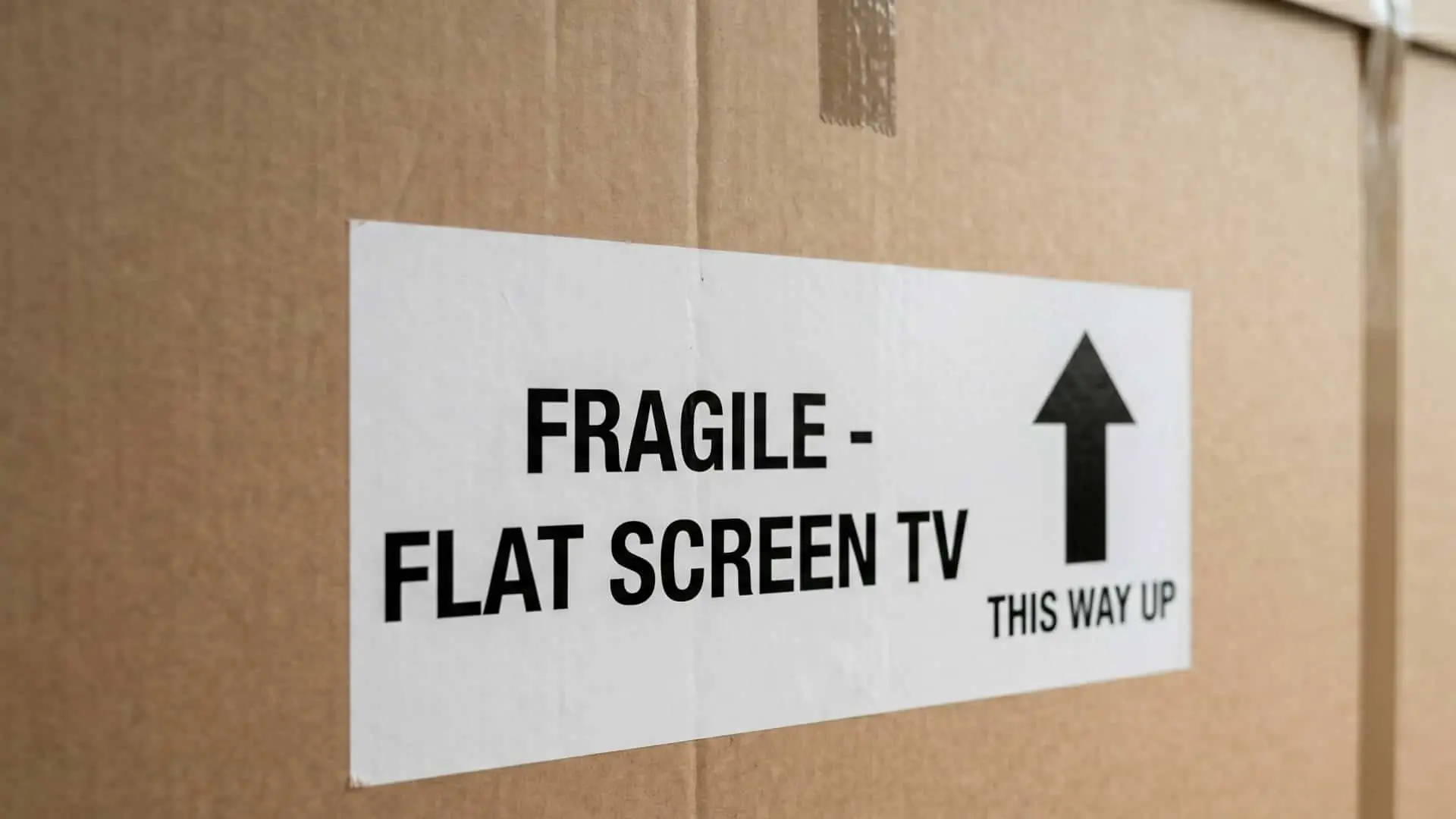 close-up of a white adhesive shipping label affixed to a brown cardboard removal box. The label features bold black text reading ‘FRAGILE - FLAT SCREEN TV’ and a clear upward-pointing arrow next to the text ‘THIS WAY UP’