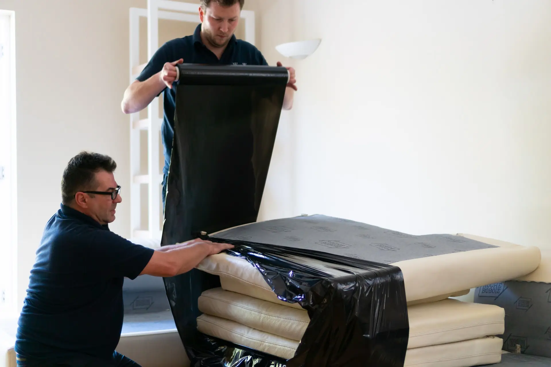 Two professional movers from W & W Movers meticulously wrapping a large, folded foam mattress in protective black stretch film, preparing it for safe, dust-free transport during a home removal.