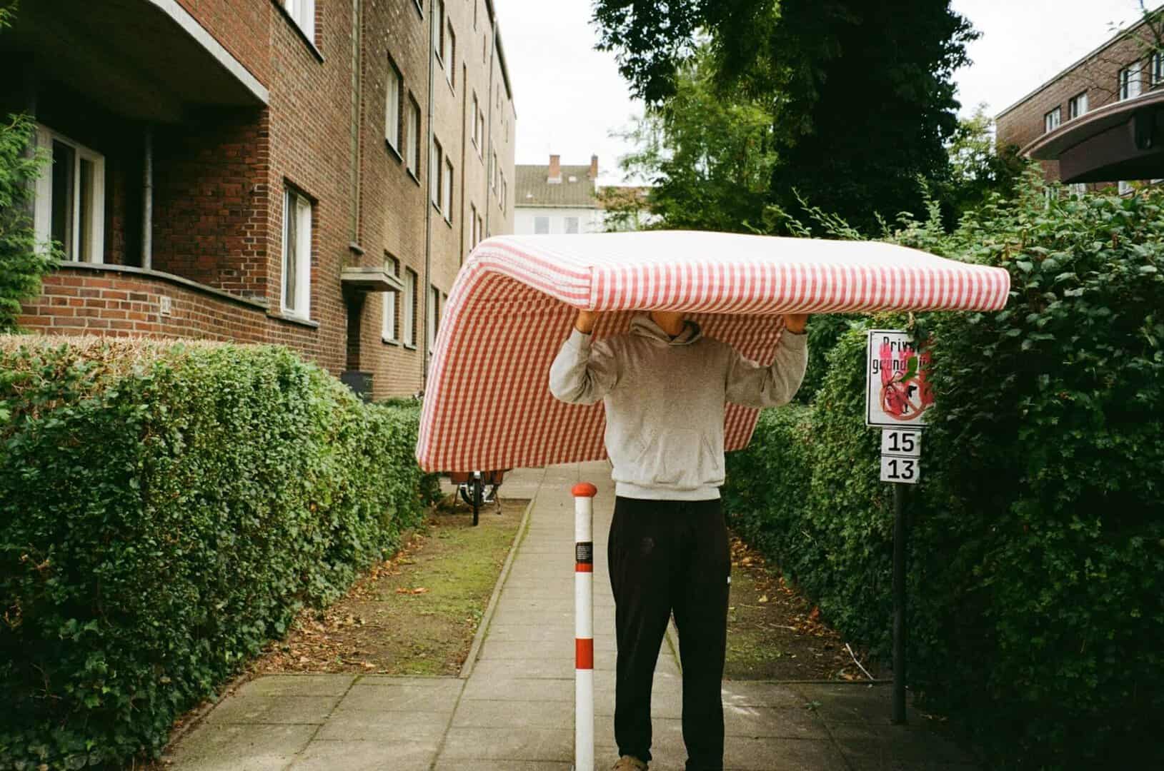 A man carrying a full-sized, non-rollable mattress, illustrating the difficulty of a solo flat-to-flat removal