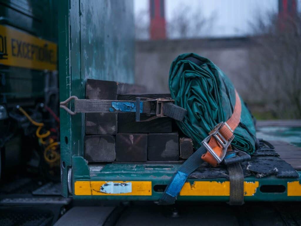 Black timber beams secured with an orange ratchet strap and a rolled green tarpaulin on a flatbed vehicle for safe transit