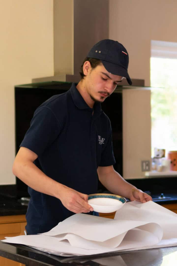A W&W Movers packer carefully wrapping a decorative china plate in tissue paper during a professional packing service at a residential property in London.